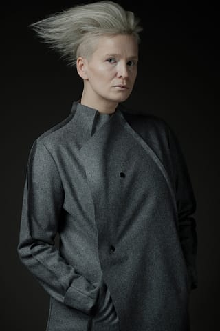 Malarski portret kobiety w trakcie sesji modowej Kobieta z krótkimi, platynowymi blond włosami ułożonymi do góry stoi z rękami w kieszeniach, ubrana w szary płaszcz. Subtelne światło na tym wyjątkowym portrecie podkreśla malarski styl fotografa.