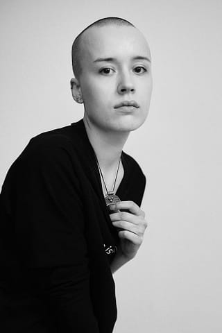 Sesja zdjęciowa czarno-białych portretów kobiety Kobieca sesja zdjęciowa. Portret młodej kobiety z ogoloną głową, ubranej w ciemną koszulę i naszyjnik, patrzącej w obiektyw z neutralnym wyrazem twarzy.