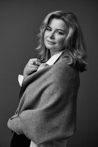 Czarno-białe zdjęcia ze studyjnej sesji kobiecej Piękno czarno-białego portretu, fotografii z sesji kobiecej, przedstawiający kobietę o falujących włosach w jasnej koszuli i udrapowanym szalu, wpatrującą się w aparat z lekkim uśmiechem.