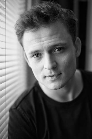 Czarno-biały headshot agencyjny do portfolio aktora Czarno-biały headshot aktorski mężczyzny z krótkimi włosami, ubranego w ciemną koszulę, patrzącego bezpośrednio w obiektyw aparatu. Portret aktora.