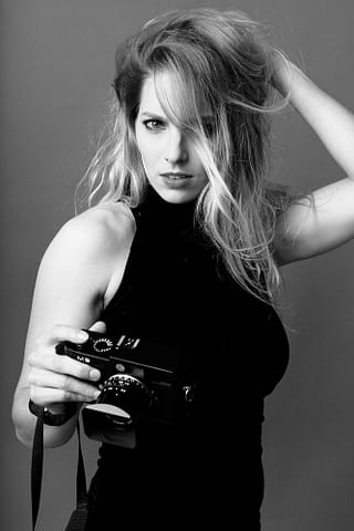 Aktorski portret kobiety w czarno-białej stylistyce Kobieta z długimi, falującymi włosami ubrana w czarny top bez rękawów trzyma aparat Leica i wpatruje się intensywnie w kamerę, jedną rękę trzymając we włosach. Ten wyrazisty, kontrastowy portret aktorki został uchwycony w czerni i bieli, ulubionej stylistyce fotografa.