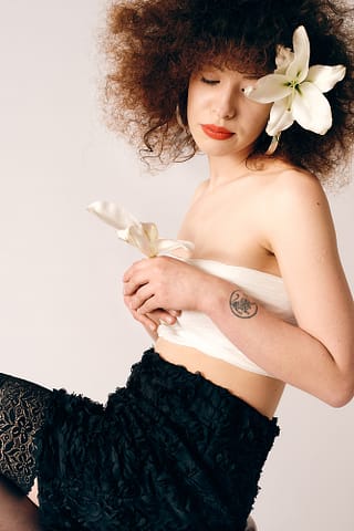 Subtelny portret z kobiecej sesji zdjęciowej w Warszawie Kobieta z kręconymi brązowymi włosami ozdobionymi dużym białym kwiatem lilii, z zamkniętymi oczami, w subtelnej kobiecej stylizacji - biały top bandeau i czarna teksturowana spódnica, czarne samonośne pończochy, biała lilia w dłoni, tatuaż widoczny na lewym przedramieniu. Sesja kobieca w warszawskim studio.