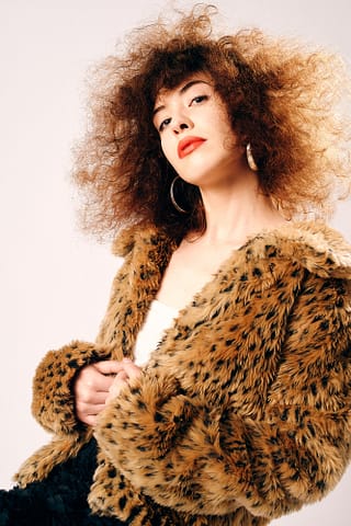 Stylizowana sesja kobieca. Sesja zdjęciowa w prezencie. Kobieta z kręconymi włosami ma na sobie sztuczne futro w panterkę i biały top, spogląda w obiektyw fotografa portrecisty, pozuje pewnie do ujęcia. Stylizowana sesja kobieca w Warszawie.