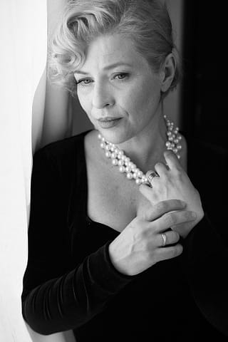 Kobieca sesja aktorska w warszawskim studio Czarno-białe zdjęcie z aktorskiej sesji zdjęciowej dojrzałej kobiety: kobieta z krótkimi, falującymi blond włosami w ciemnej sukience, naszyjniku z pereł i pierścionkach stoi przy oknie w delikatnym świetle.