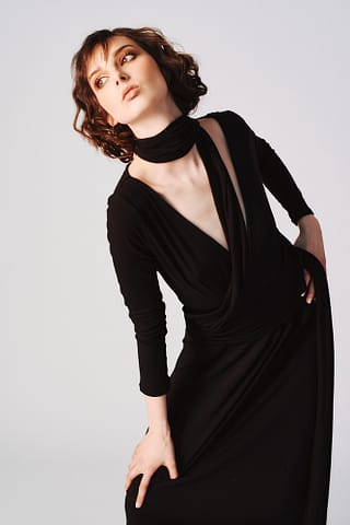 Klasyczna sesja zdjęciowa mody w Warszawie Kobieta z krótkimi, falującymi brązowymi włosami ma na sobie czarną sukienkę z długimi rękawami i głębokim dekoltem w szpic oraz pasujący szal. Pozuje pewnie, zgodnie z instrukcjami fotografa - specjalisty w dziedzinie mody.