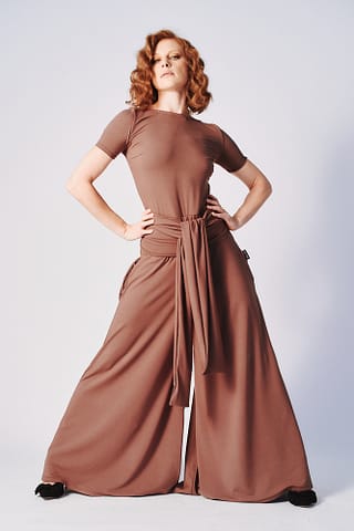 Klasyczna fotografia modowa Kobieta z falistymi rudymi włosami ubrana w jasnobrązowy kombinezon z krótkim rękawem, szerokimi nogawkami i długim wiązaniem w talii, stoi z dłońmi opartymi o biodra w klasycznej pozie fotografii modowej. Zdjęcie studyjne.