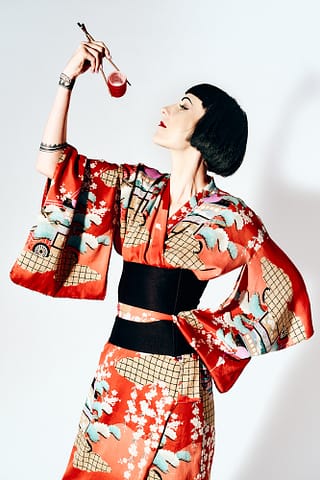 Stylizowana kobieca sesja zdjęciowa w studio Kobiecy portret w japońskiej stylistyce: kobieta w czerwonym kwiecistym kimonie z czarnym obi trzyma uniesionymi pałeczkami wałek do włosów jak porcję sushi, jej czarne włosy i barwny strój kontrastują z jasnym tłem. Studyjna sesja zdjęciowa w Warszawie.