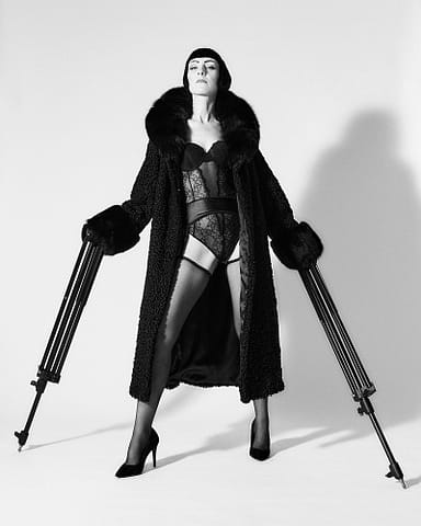 Odważna kobieca sesja zdjęciowa w Warszawie Czarno-biały portret kobiety w bieliźnie, wysokich obcasach i teksturowanym płaszczu z futrzanymi mankietami, modelka stoi pewnie, używając statywów fotograficznych jako lasek. Odważny, dramatyczny obraz podkreśla jej silną osobowość.