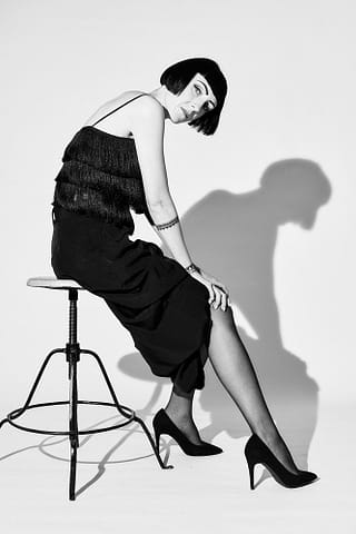 Czarno-biały portret kobiecy w stylizowanej sesji zdjęciowej Kobieta z krótką fryzurą ubrana w czarny top bez rękawów z frędzlami, czarną spódnicę i wysokie obcasy siedzi bokiem na wysokim stołku na jednolitym tle - przykład klasycznej kontrastowej fotografii kobiecej - z wyraźnym cieniem rzucanym na ścianę.
