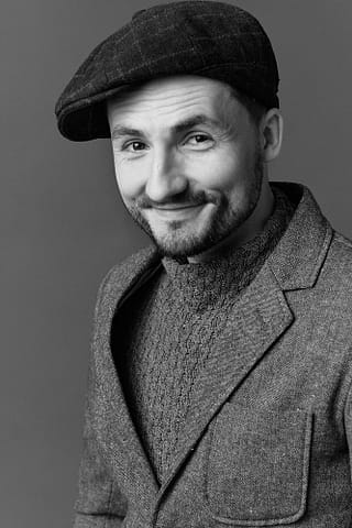 Czarno-biały portret do portfolio aktora Czarno-biały portret uśmiechniętego aktora w kaszkiecie, teksturowanym swetrze i marynarce, z krótkimi włosami i przystrzyżoną brodą, sesja zdjęciowa w klimacie warszawskiej Pragi - idealna inspiracja do aktorskiej sesji portretowej w Warszawie.