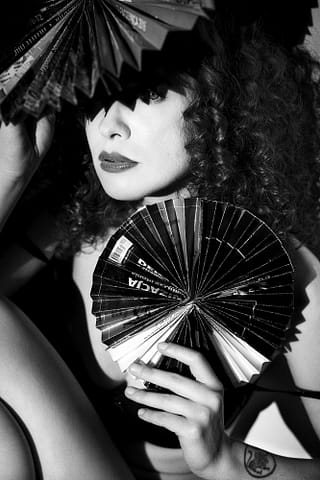 Stylizowana sesja kobieca w Warszawie Czarno-biały stylizowany portret kobiety z kręconymi włosami trzymającej dwa papierowe wachlarze - jeden nad głową rzucający cień na oczy, drugi przed klatką piersiową. Nosi ciemną szminkę i ma tatuaż na ramieniu. Sesja studyjna.