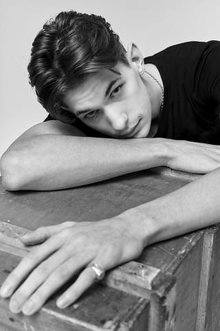 Portret w trakcie sesji polaroidów początkującego modela Czarno-biały portret młodego mężczyzny z ciemnymi włosami, opierającego głowę i ramiona na drewnianym pudełku. Patrzy prosto w obiektyw z neutralnym wyrazem twarzy, ma na sobie ciemny t-shirt i sygnet na palcu.