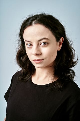 Zdjęcia agencyjne do aktorskiego portfolio Kobieta o falujących ciemnych włosach i jasnej karnacji ubrana w czarny t-shirt stoi na jednolitym jasnoniebieskim tle, patrząc spokojnie w kamerę, sesja zdjęciowa headshotów do agencji aktorskiej.