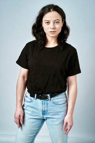 Zdjęcia studyjne do agencji aktorskich Kobieta z falującymi ciemnymi włosami ubrana w czarną koszulkę, jasnoniebieskie dżinsy i czarny pasek stoi na prostym jasnoszarym tle, patrząc pewnie w kamerę - tak właśnie powstają sprawdzone kadry zdjęć do agencji aktorskiej.