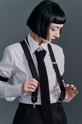 Studyjna fotografia produktowa mody Kobieta o krótkich czarnych włosach ma na sobie białą koszulę, czarne szelki i czarny krawat, patrząc w dół, spogląda w kierunku dłoni trzymającej szelki. Czerwone paznokcie dodają kontrastu. Czyste spojrzenie na fotografię produktową mody.