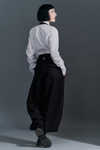 Szlachetna fotografia modowa Kobieta z krótką czarną fryzurą bob, ubrana w białą koszulę z długim rękawem i luźne czarne spodnie z wysokim stanem, stoi odwrócona przodem do ciemnoszarego tła, zdjęcie podkreśla piękno prostych fotograficznych ujęć.