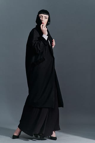 FIlmowe fotografie kobiet w stylizacjach modowych Kobieta o charyzmatycznej urodzie ubrana w długi czarny płaszcz i szerokie czarne spodnie trzyma wysoko postawiony kołnierz, stylizowana poza podkreśla filmowy charakter zdjęć.