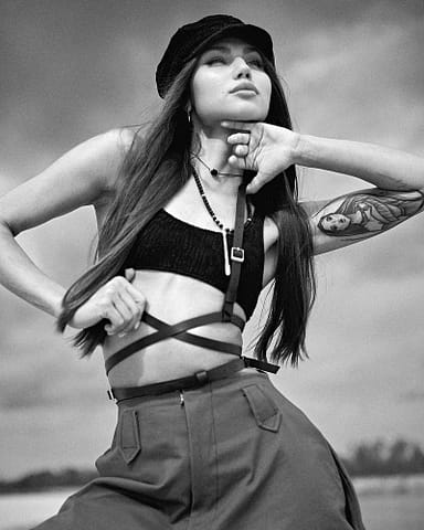 Sesja czarno-białych zdjęć modowych Kobieta pozuje na zewnątrz w czarnym crop topie, spodniach z wysokim stanem i czapce. Ma długie włosy, naszyjnik z paciorkami i tatuaż na ramieniu. Czarno-białe zdjęcie oddaje najlepszy klimat w fotografowaniu mody z dramatycznym, stylowym charakterem.