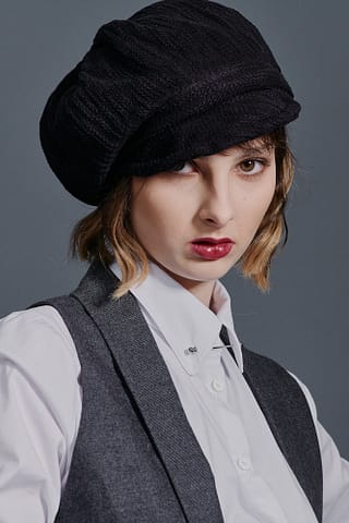 Portret i moda na jednej fotografii Kobieta z falującymi jasnobrązowymi włosami ma na sobie czarny kaszkiet, białą koszulę zapinaną na guziki i szarą kamizelkę, patrzy bezpośrednio w kamerę z poważnym wyrazem twarzy. Kobiecy portret zrealizowany w trakcie modowej sesji zdjęciowej.