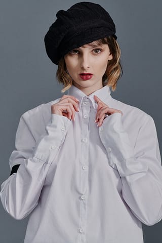 Produktowa sesja zdjęciowa mody Kobieta ubrana w białą koszulę zapinaną na guziki i czarny kaszkiet stoi na szarym tle, patrząc w kamerę z rękami przy kołnierzu - elegancki przykład fotografii ubrań w studio.