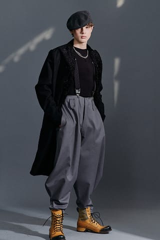 Modowa sesja zdjęciowa produktów Chłopak ubrany w szary kaszkiet, czarny płaszcz, czarną koszulę, szare szerokie spodnie z szelkami i żółte sznurowane buty stoi na szarym tle - fotografia mody z charakterem.