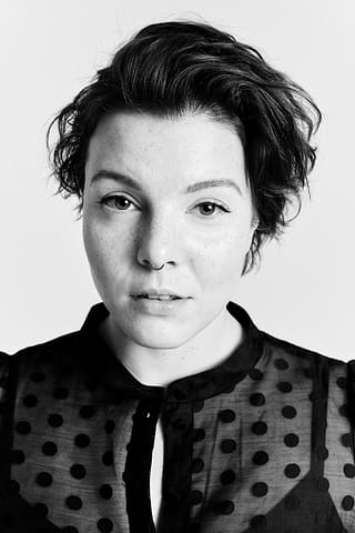 Czarno-biały portret wokalistki muzyka Czarno-biała fotografia wokalistki z krótkimi, falowanymi włosami w przezroczystej bluzce w kropki, patrzącej neutralnie prosto w obiektyw na jednolitym tle w studio.