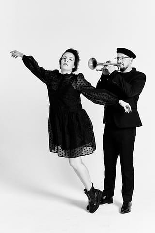 Fotografia muzyczna duetu artystów Kobieta w czarnej sukience i butach pozuje z wyciągniętymi ramionami, podczas gdy mężczyzna w czarnym stroju i berecie gra na trąbce tuż za nią; oboje znajdują się na białym tle podczas studyjnej sesji zdjęciowej tego muzycznego duetu.