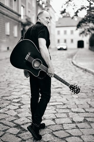 Zdjęcie na okładkę albumu muzyka Mężczyzna w ciemnym ubraniu idzie brukowaną ulicą, trzymając przy boku gitarę akustyczną i oglądając się przez ramię - sugestywny, czarno-biały portret na okładkę płyty, z budynkami i samochodem w tle.