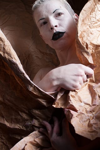 Stylizowana kobieca sesja zdjęciowa Kobieta z krótkimi blond włosami pozuje do fotograficznego portretu w teatralnym makijażu - ma ciało pomalowane na biało, usta umalowane czarną szminką i narysowane linie brwi - zawinięta jest w pakowy, pomięty brązowy papier, patrzy w górę z dramatycznym wyrazem twarzy.