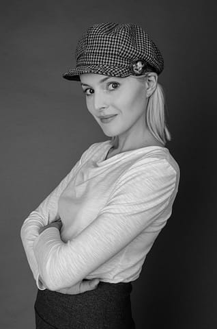 Portret kobiety w trakcie studyjnej sesji zdjęciowej Czarno-białe zdjęcie z kobiecej sesji modowej, przedstawiające kobietę o jasnych włosach w czapce w pepitkę i jasnej koszuli z długim rękawem, stojącą w studio ze skrzyżowanymi ramionami i lekko uśmiechniętą.