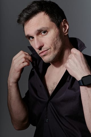 Portret aktora do portfolio Mężczyzna w ciemnej koszuli poprawia kołnierzyk, patrząc pewnie w kamerę, stojąc na szarym tle. Ten wyjątkowy portret aktorski oddaje jego silny charakter.