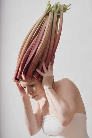 Stylizowana kobieca sesja zdjęciowa Stylizowany portret kobiety przedstawia osobę w białym topie bez ramiączek, stojącą na naturalnym tle w koronie z kilkunastu długich łodyg rabarbaru na głowie jak wysokim kapeluszu.