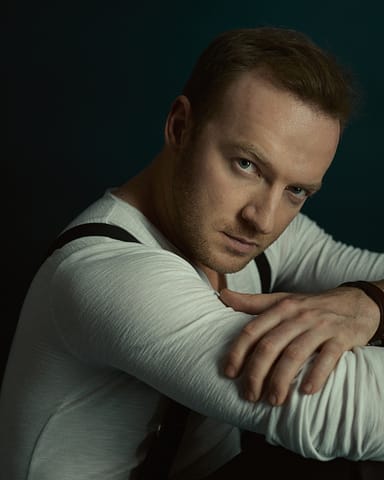 Studyjny portret aktora w modowej stylizacji Mężczyzna z krótkimi jasnobrązowymi włosami, ubrany w białą koszulę z długim rękawem i czarne szelki, siedzi opierając ręce na kolanach, wpatrując się uważnie w kamerę w tym studyjnym portrecie aktorskim na ciemnym tle.