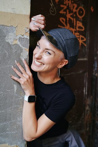 Modowa sesja zdjęciowa w Warszawie Wesoła kobieta z krótkimi włosami, w kaszkiecie, dużych kolczykach, czarnej koszulce, uśmiecha się szeroko i opiera o zniszczoną ścianę z graffiti w tle - żywy kobiecy portret pełen osobowości.