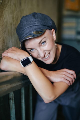 Portret kobiety w modowej miejskiej stylizacji Uśmiechnięta kobieta w szarym kaszkiecie, czarnej koszulce i smartwatchu opiera ręce i głowę o zieloną barierkę, patrząc w kamerę podczas sesji zdjęciowej na ulicach Warszawy.