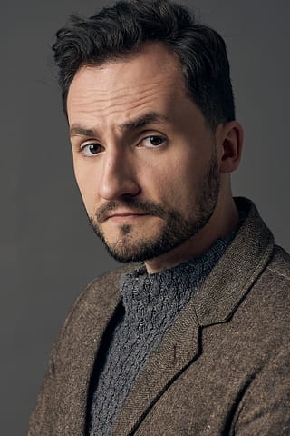 Headshoty do portfolio aktora Mężczyzna z krótkimi ciemnymi włosami i przystrzyżoną brodą nosi brązową kurtkę na teksturowanym szarym swetrze, patrząc poważnie w kamerę na prostym szarym tle na tym profesjonalnym zdjęciu aktorskim.