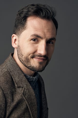 Headshoty do portfolio aktora Mężczyzna z krótkimi ciemnymi włosami i przystrzyżoną brodą nosi brązową teksturowaną marynarkę na szarym swetrze, pozując na prostym szarym tle i lekko uśmiechając się do aparatu - idealny portret do aktorskiego portfolio.