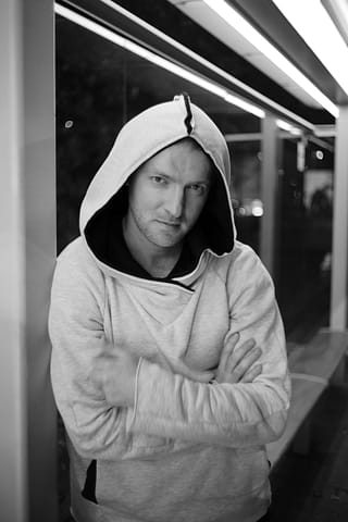 Sesja zdjęciowa w modowej miejskiej stylizacji Mężczyzna w bluzie z kapturem stoi ze skrzyżowanymi rękami, opierając się o ścianę w nocy. To czarno-białe ujęcie oddaje klimat miejskiej sesji zdjęciowej mody, z jasnymi neonowymi światłami w tle.