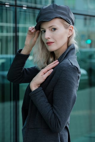 Miejska modowa sesja zdjęciowa na ulicach Warszawy Kobieta o blond włosach w ciemnej czapce i szarej marynarce pozuje pewnie na zewnątrz, dotykając daszka kaszkietu podczas miejskiej modowej sesji zdjęciowej. Tło przedstawia szklany budynek z odbiciami i rozmytą scenę uliczną.