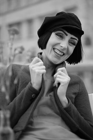 Kobiecy portret z modowej sesji miejskiej Kobieta z krótką fryzurą bob w kaszkiecie uśmiecha się szeroko, ciągnąc za kołnierz płaszcza. Czarno-białe zdjęcie doskonale oddaje kobiecą miejską stylizację modową na miękko rozmytym tle.