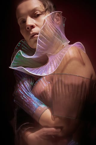Stylizowana kobieca sesja zdjęciowa Kobieta pozuje do stylizowanej sesji zdjęciowej na ciemnym tle, ubrana w półprzezroczystą, opalizującą, plisowaną szatę z dramatycznymi falbanami wokół kołnierza i ramion, tworząc eteryczny i futurystyczny wygląd.
