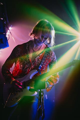 Relacja z występu live Zdjęcie z koncertu przedstawia gitarzystę z włosami do ramion, który gra na gitarze elektrycznej na scenie, oświetlonej kolorowymi zielonymi i żółtymi światłami scenicznymi. Ma na sobie wzorzystą koszulę i skupiony wyraz twarzy.