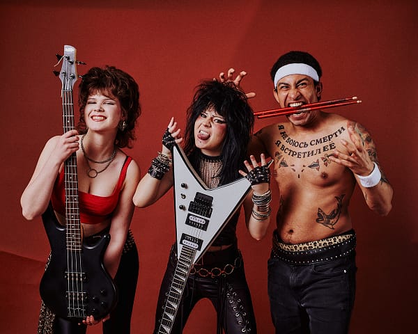Sesja zdjęciowa zespołu glam metalowego Warszawa Troje energicznych ludzi w strojach rockowych pozuje z instrumentami na czerwonym tle podczas sesji zdjęciowej zespołu glam metalowego. Jedna z dziewczyn trzyma gitarę basową, druga gitarę elektryczną, a pozbawiony koszulki członek zespołu ma pałeczki perkusyjne w ustach i tatuaże na klatce piersiowej.