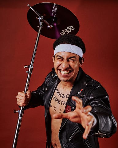 Sesja zdjęciowa muzyka Warszawa Mężczyzna w czarnej skórzanej kurtce i białej opasce na głowie uśmiecha się intensywnie, trzymając stojak na talerze podczas sesji zdjęciowej. Ma tatuaże na klatce piersiowej i pozuje z wyciągniętą ręką na żywym czerwonym tle, oddając ducha rockowej muzyki.