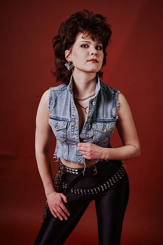 Sesja zdjęciowa zespołu muzycznego Kobieta z fryzurą mullet ma na sobie dżinsową kamizelkę bez rękawów, czarne błyszczące spodnie z paskiem nabijanym ćwiekami, odważną biżuterię i dramatyczny makijaż, pozując pewnie na bordowym tle w inspirowanej rockiem z lat 80-tych sesji portretowej artystki.