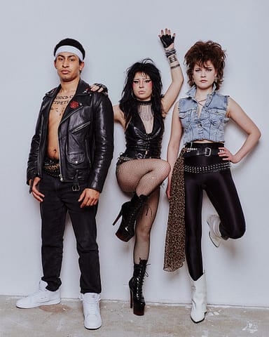 Sesja zdjęciowa zespołu muzycznego Trzy osoby w ostrych glamrockowych strojach z lat 80. pozują na tle białej ściany do sesji zdjęciowej zespołu muzycznego, ubrane w skóry, kabaretki, ćwieki i odważne akcesoria, przyjmując pewne siebie i buntownicze postawy.