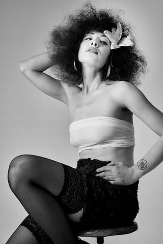 Kobieca sesja portretowa w Warszawie Kobieta z kręconymi włosami i dużym kwiatowym dodatkiem pozuje na stołku w tej efektownej czarno-białej sesji kobiecej. Ma na sobie top bez ramiączek, koronkowe szorty i pończochy, jedną rękę trzyma na głowie, a drugą na biodrze.